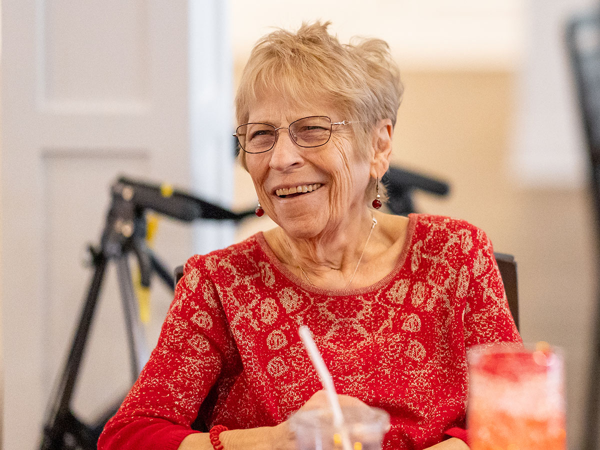 Smiling older woman with glasses sitting at table in community, wearing red blouse.