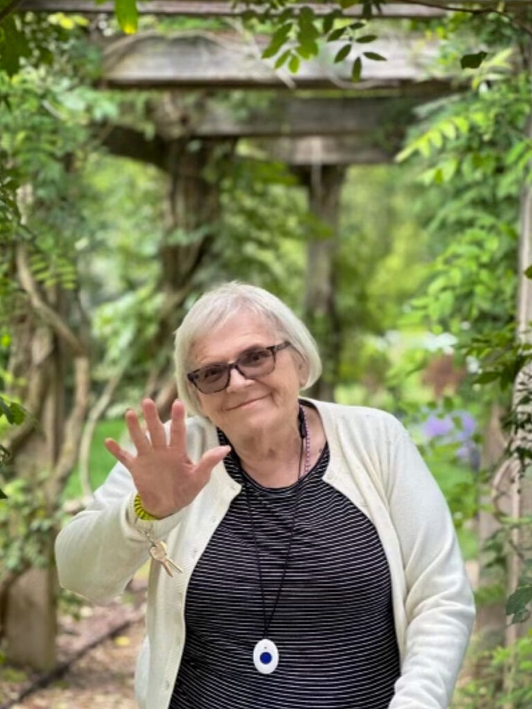 This is a current photo of Carol in her glasses with a black stripped top standing in a garden.