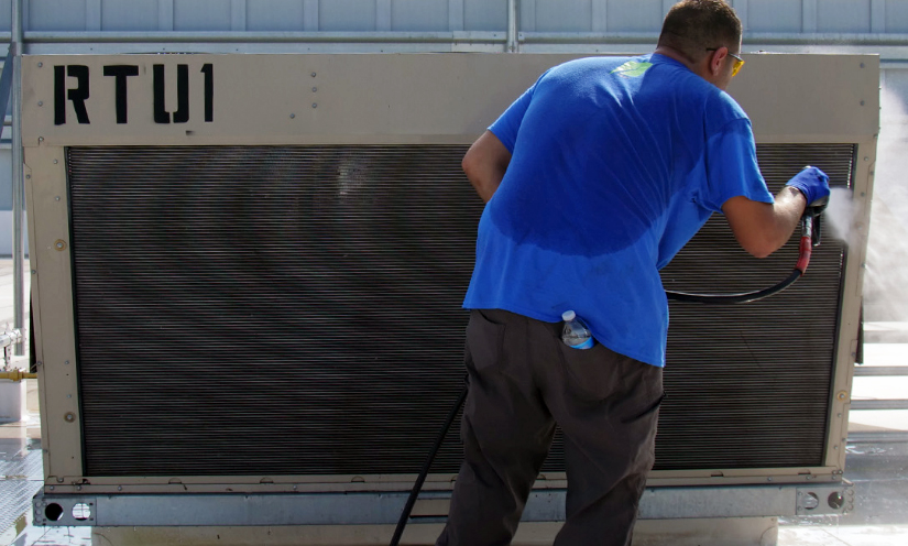 A man in a blue shirt spraying water on a large outdoor AC unit.