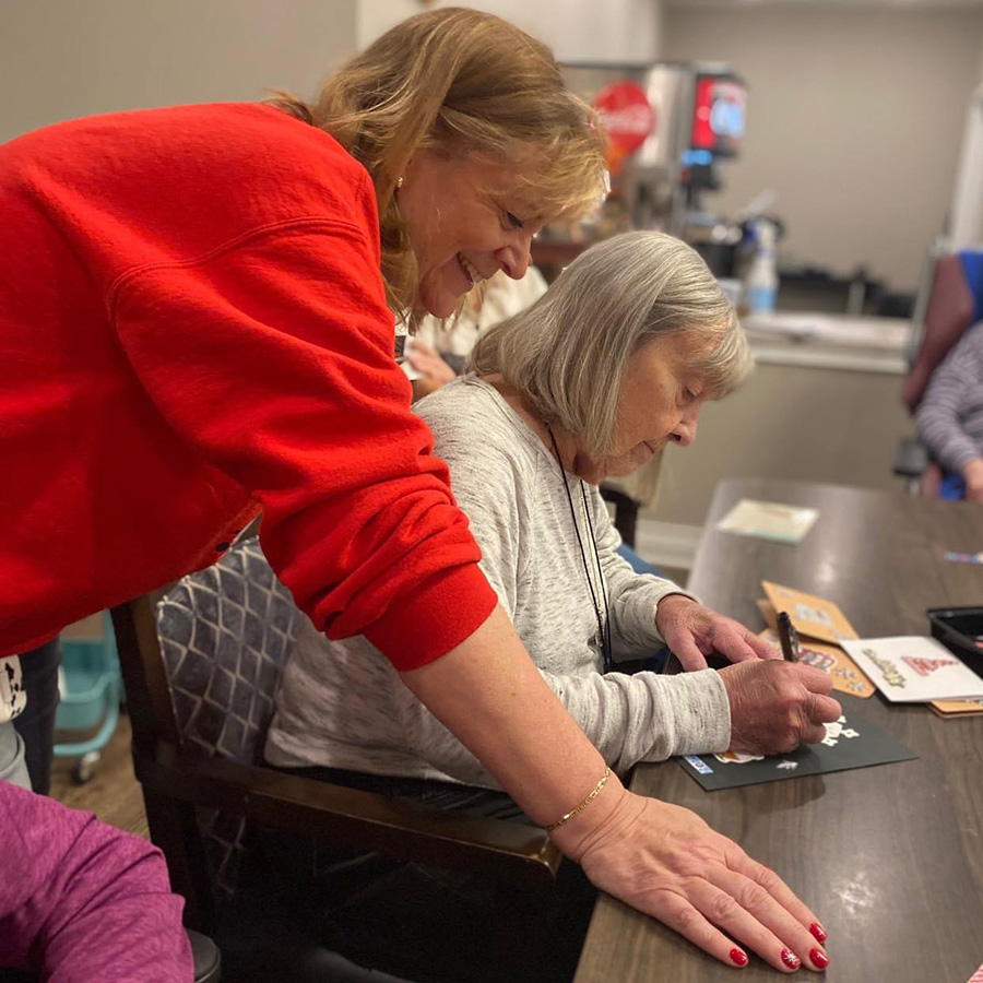 A caring volunteer assists an elderly woman in signing a card, spreading joy during the holiday season at a memory care neighborhood.