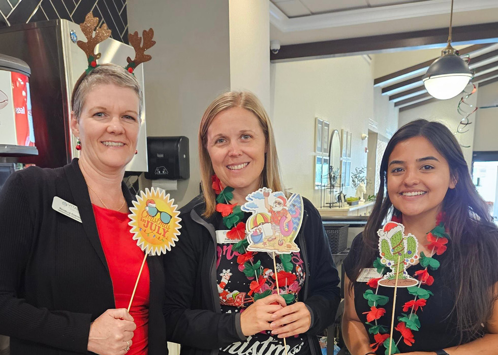 Three women dressed in festive attire hold holiday-themed props, including a 'Christmas in July' sign, standing together and smiling in a brightly lit room.