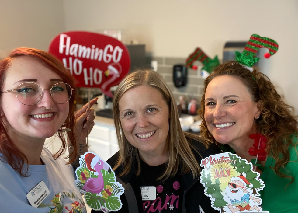 Three smiling women hold holiday-themed props, including a 'FlaminGO Ho Ho Ho' sign and 'Christmas in July' decorations, celebrating in a festive environment.