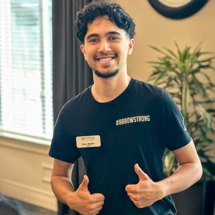 A staff member at Vitalia Senior Living smiles and gives two thumbs up while wearing a name tag and a shirt with the text '#ArrowStrong.