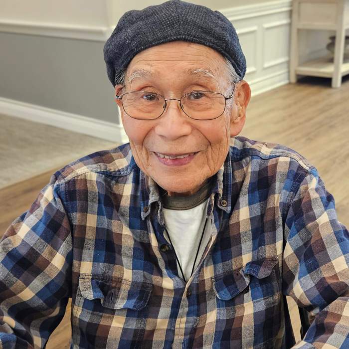 A senior resident smiles warmly, wearing a checkered shirt and glasses, while seated indoors.