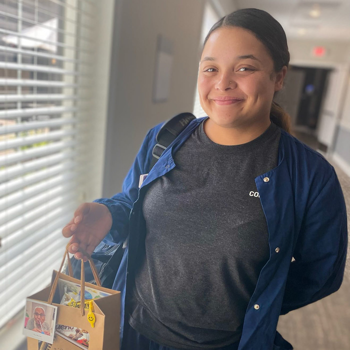 A staff member smiles while holding a small gift bag, dressed casually in a dark shirt and blue jacket.