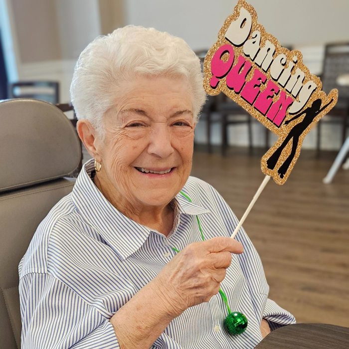 A smiling senior resident holds up a sign that reads 'Dancing Queen' during a fun event in the senior living community.