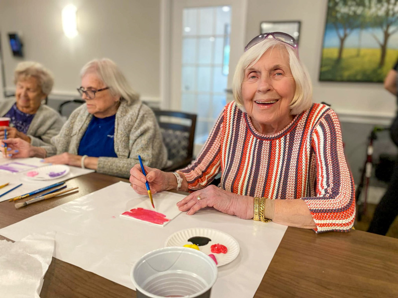 A senior resident participates in a painting activity, smiling while holding a brush. Other residents are seated nearby, also engaged in painting.