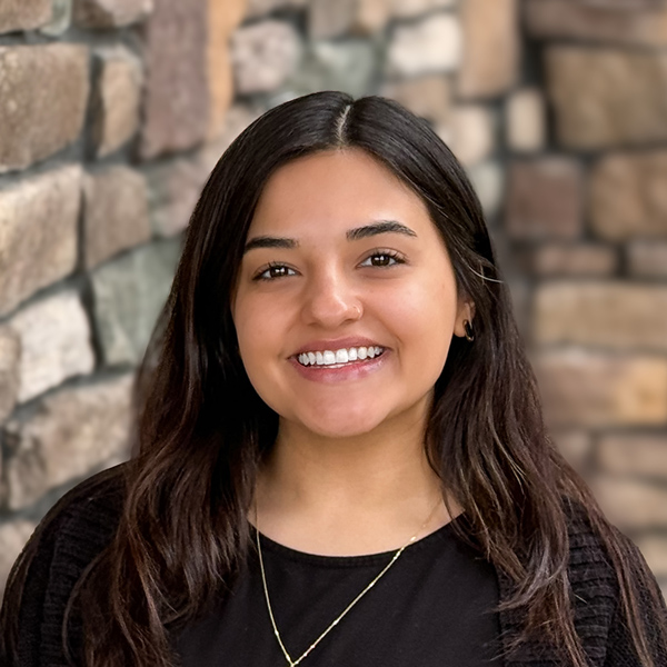 Lauren Garcia, Senior Living Director at Vitalia North Olmsted, smiling outside in front of a multi-colored stone wall while wearing a black top and gold necklace.