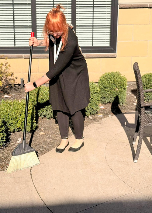 Employee sweeping the outdoor patio walkway in bright sunlight at a senior living community.
