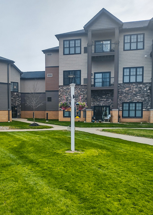 Well-maintained green lawn and walking path in front of a senior living apartment building, featuring hanging flower baskets on a white post.