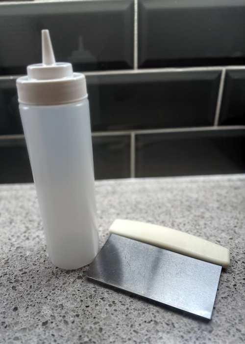 Plastic squeeze bottle, metal bench scraper, and white dough scraper placed on a speckled kitchen counter, highlighting versatile and underrated kitchen tools.