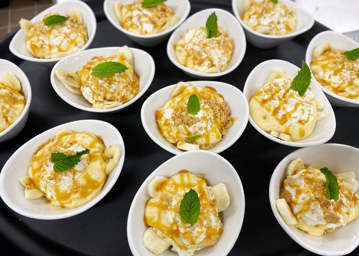 Mini banana pudding bowls topped with whipped cream, caramel drizzle, crushed cookies, and fresh mint, arranged on a black serving tray.