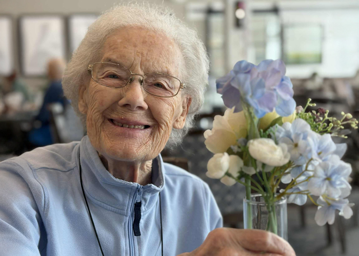 Smiling senior woman in a light blue zip-up jacket holding a small bouquet of pastel flowers in a vase