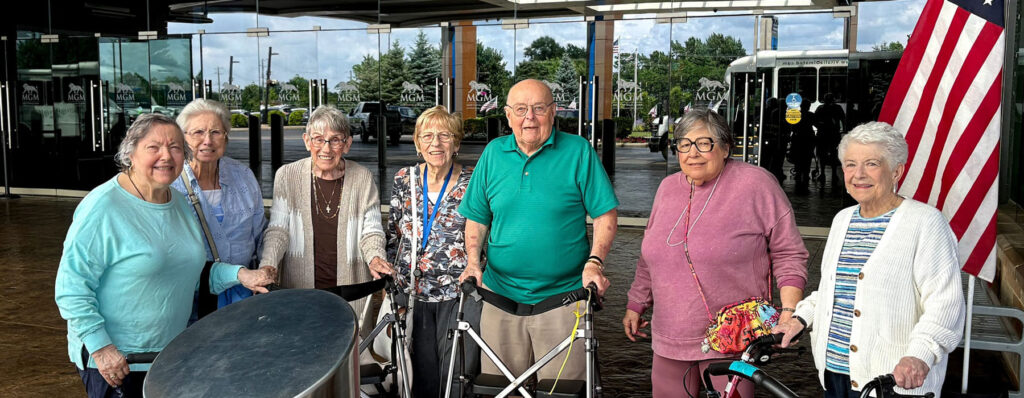 Group of smiling senior residents standing outside MGM Casino, some using walkers, enjoying a day trip from their senior living community.