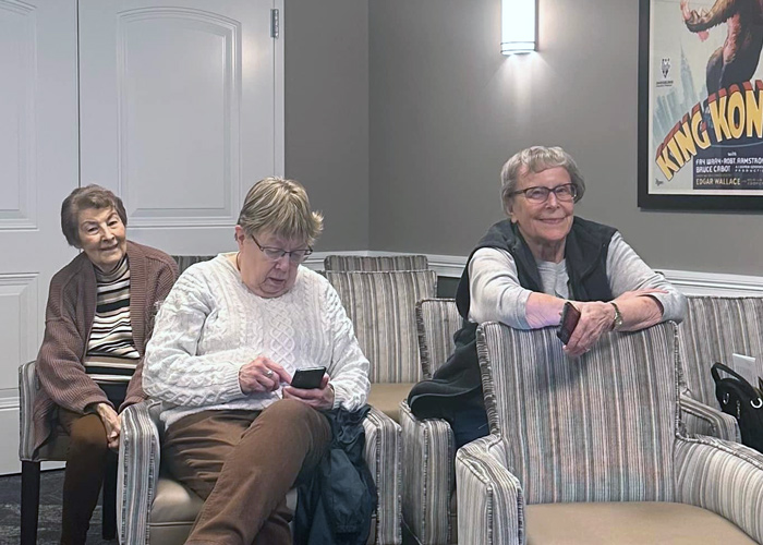 Three senior residents seated in striped chairs, smiling and using smartphones during a tech session.
