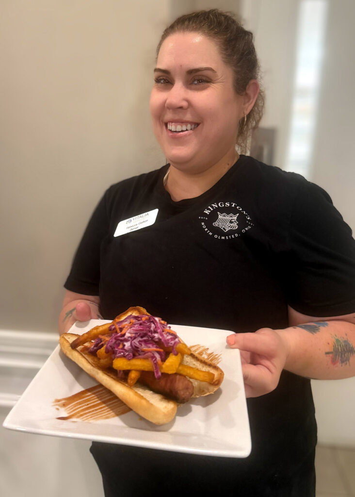 Team member at Vitalia North Olmsted smiles while holding a gourmet sausage sandwich topped with crispy fries and colorful slaw on a toasted bun, ready to serve.