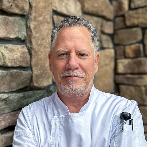 David Romano, Culinary Director at Vitalia North Olmsted, wearing a white chef coat and standing in front of a stone wall.