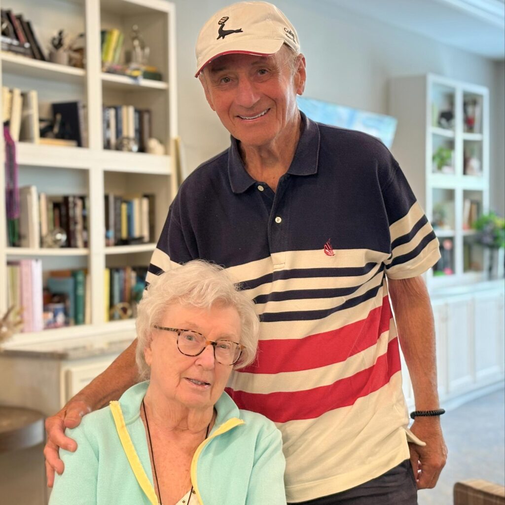 An older man in a striped polo and white cap stands behind a smiling senior woman seated in front of a bookcase, both enjoying time together at the community.