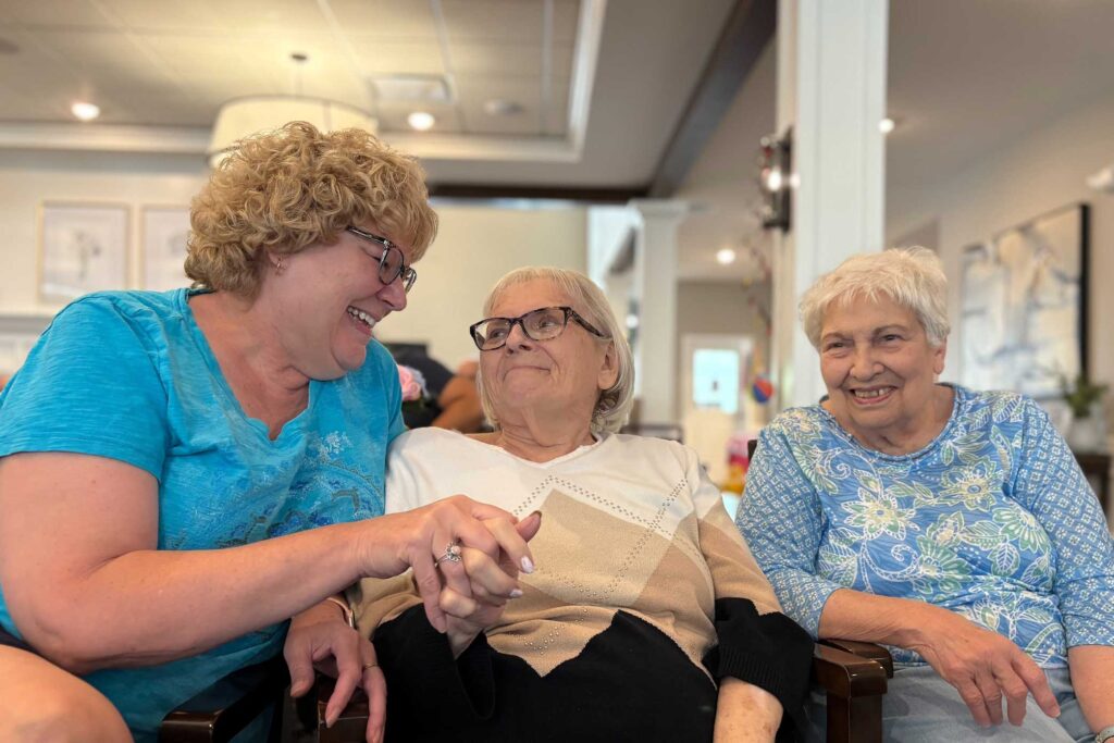 A woman in a bright blue shirt laughs and holds hands with a smiling senior resident, while another woman in a floral blue top joins in the cheerful moment together.