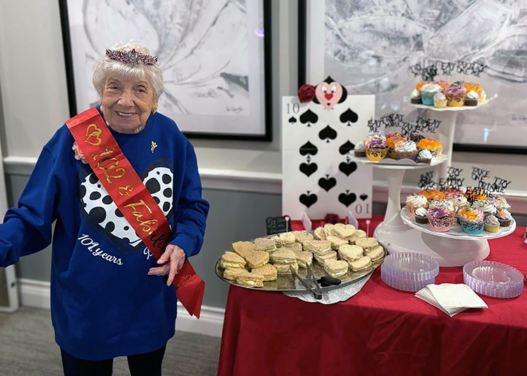 A resident celebrates her 102nd birthday with a crown and sash, smiling next to a themed table filled with treats.
