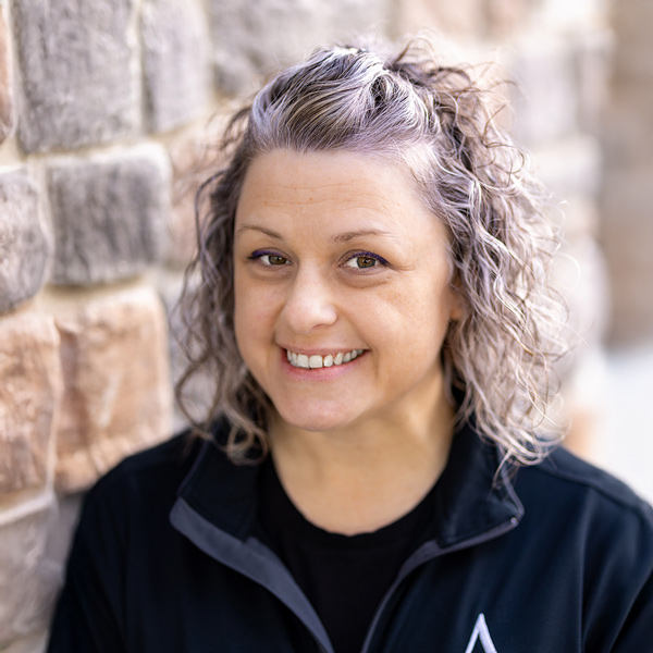 Ashley Bago, Wellness Director at Vitalia North Olmsted, smiling in a professional headshot with curly silver hair, wearing a black jacket, posed against a softly blurred stone wall background.