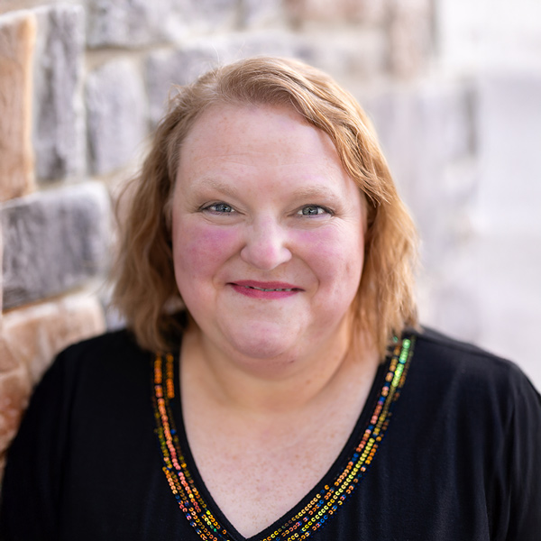 Becky Gibson, Resident Services Director at Vitalia North Olmsted, smiling in a professional headshot, wearing a black top with colorful beaded neckline against a softly blurred stone wall background.