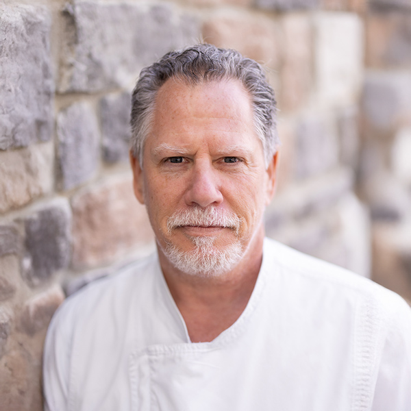 David Romano, Culinary Director at Vitalia North Olmsted, in a professional headshot wearing a white chef coat, standing against a softly blurred stone wall background.