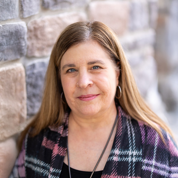 Gretchen Banak, Business Office Director at Vitalia North Olmsted, in a professional headshot with long light brown hair, wearing a patterned cardigan, standing against a softly blurred stone wall background.