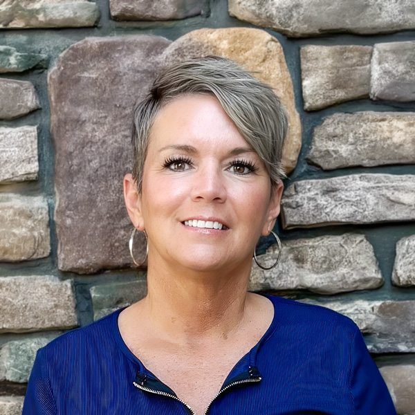 Tracy Barbee, Senior Living Director at Vitalia North Olmsted, looking at the camera in a professional headshot with short silver pixie-cut hair, hoop earrings, a royal blue blouse, and a stacked stone wall background.